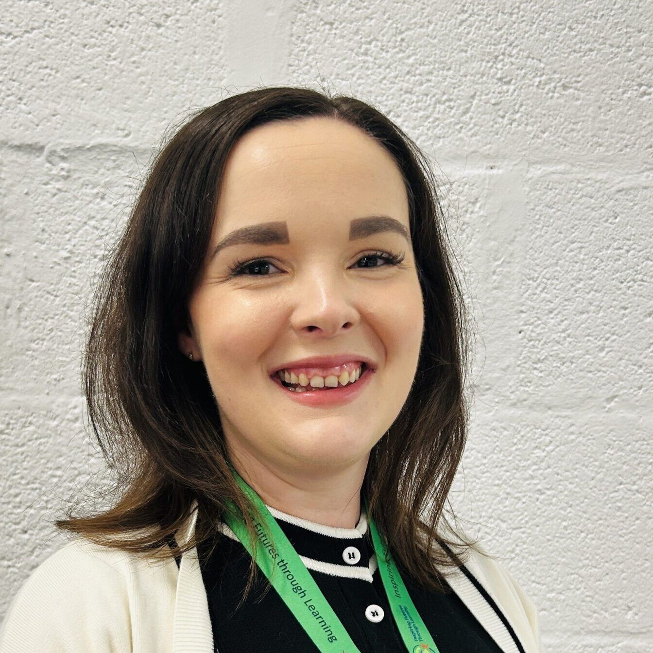 Young woman smiling at the camera, wearing a green lanyard, standing against a white brick wall.